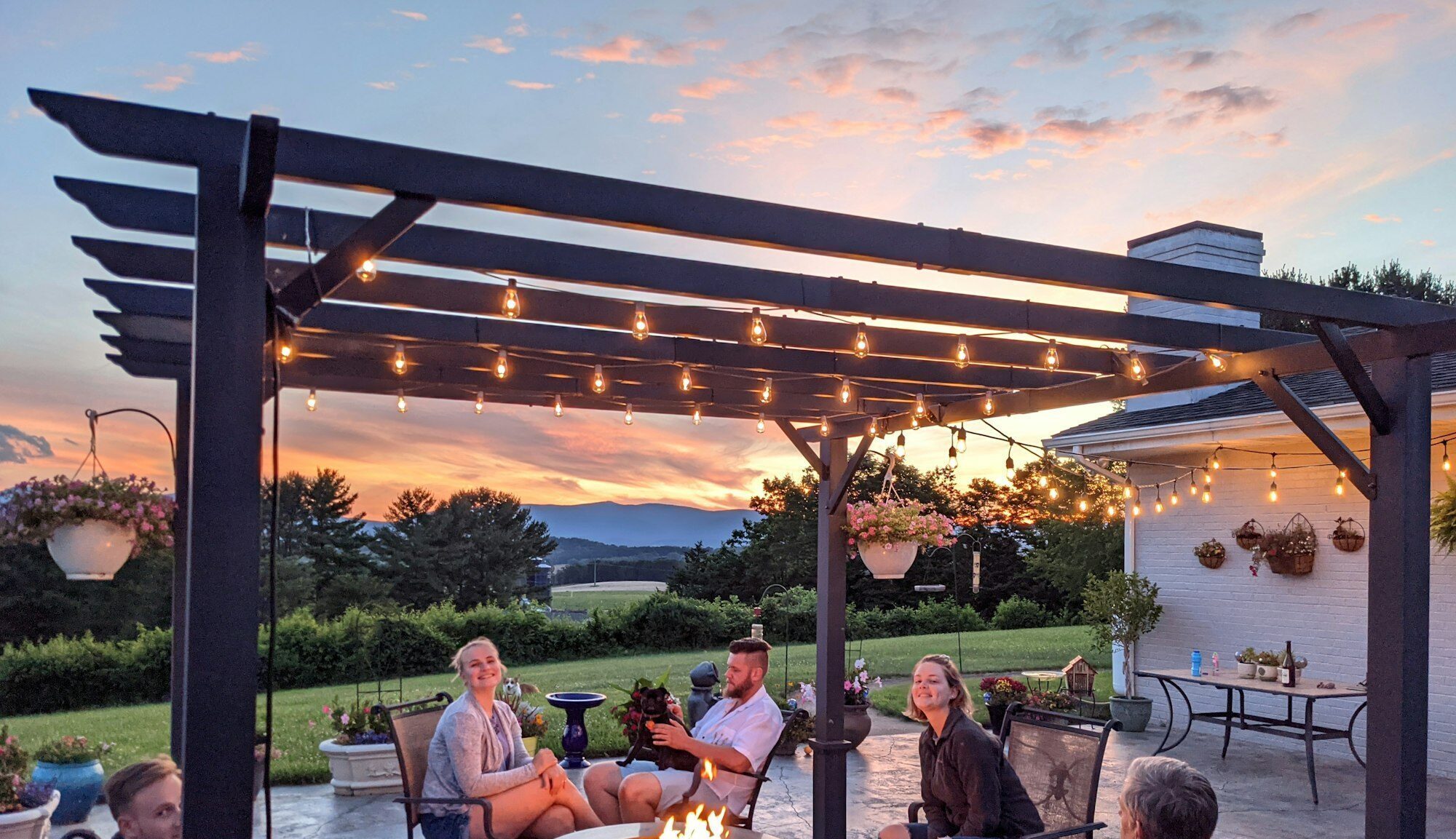 A family relaxing around a fire table in outdoor seating area under pergola with lights at sunset